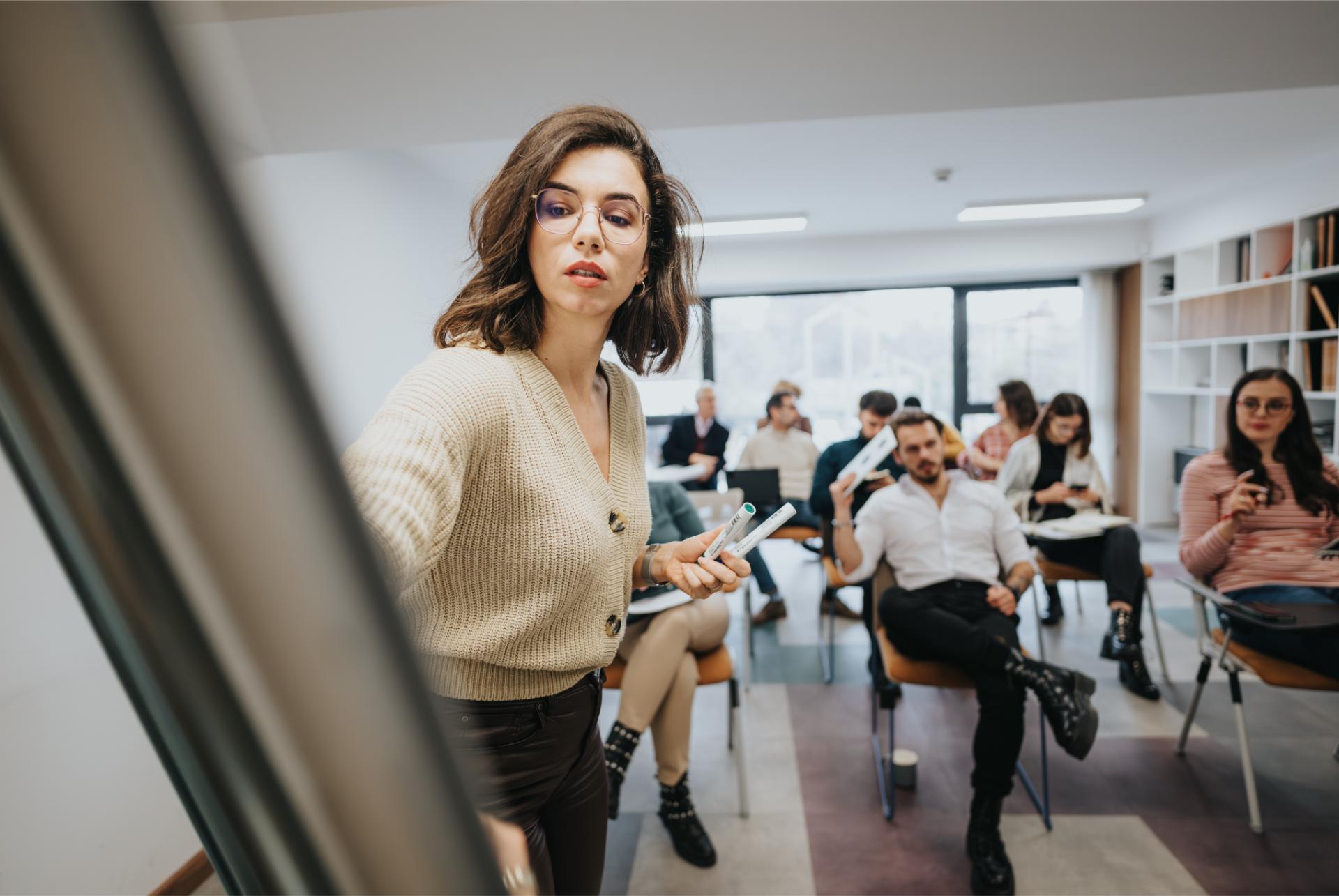 A young woman leads a presentation at a business workshop confidently engaging with a group of professionals seated in a stylish office setting
