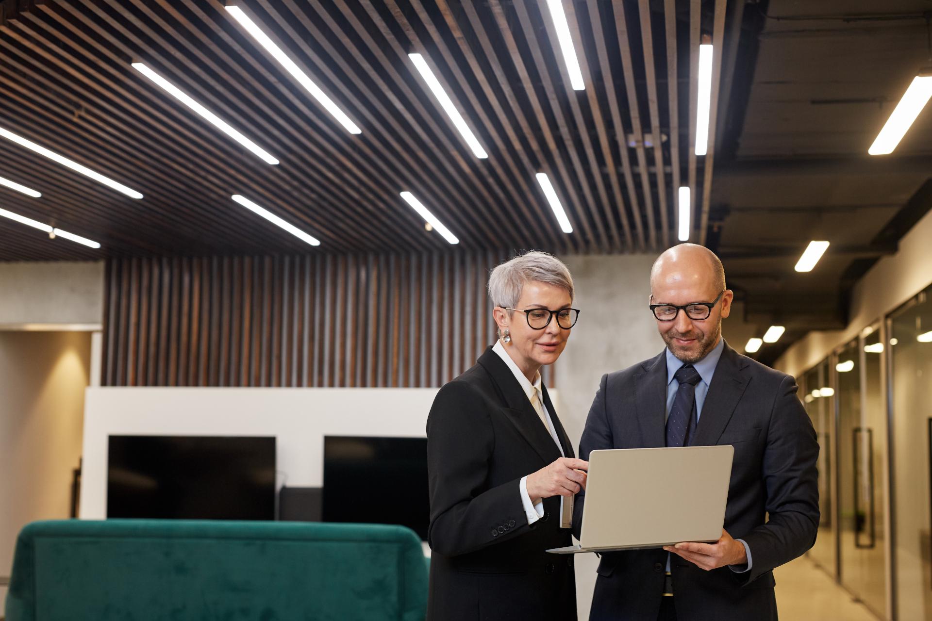 Waist up portrait of two mature business people holding laptop while standing in modern office interior copy space