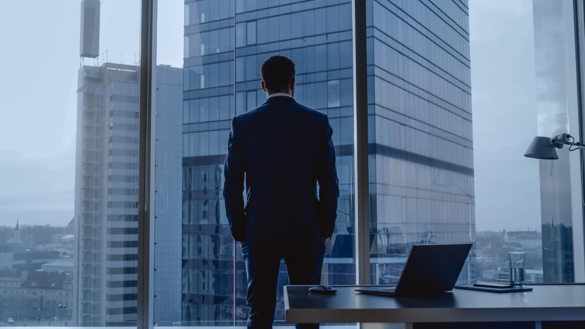 Back View of the Thoughtful Businessman wearing a Suit Standing in His Office Hands in Pockets Looking out of the Window