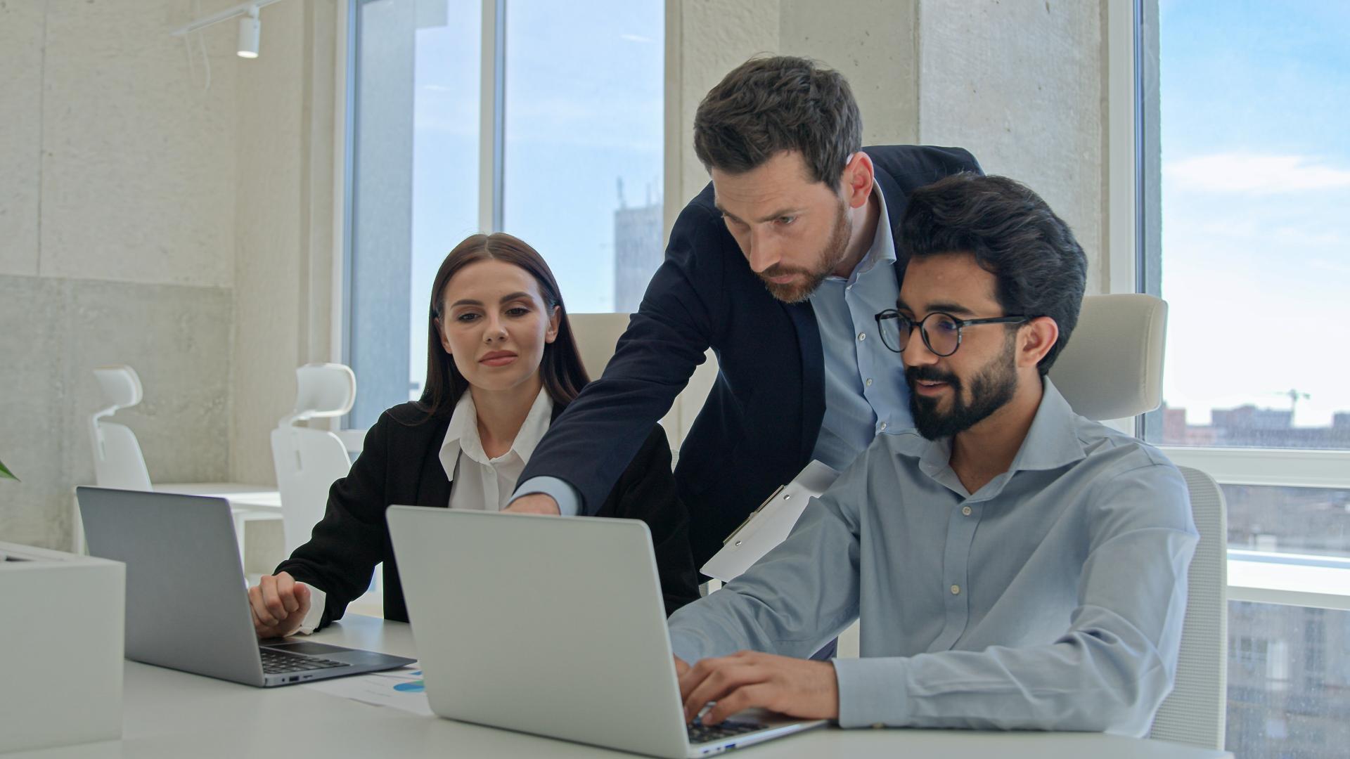 Male CEO boss leader man businessman help office coworkers with computer work check online project on laptop talking to diverse multiracial business team colleagues brainstorm analyze sales paperwork